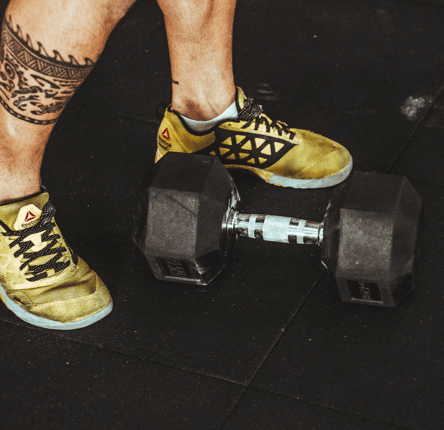 Eine Person in gelben Sportschuhen mit einer geometrischen Tätowierung auf dem Bein steht neben einer schweren Hantel auf dem Boden eines schwarzen Fitnessstudios.