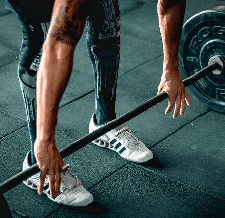 Eine Person in Sportkleidung und Gewichtheberschuhen bereitet sich darauf vor, in einem Fitnessstudio eine Langhantel mit Gewichten zu heben. Nur ihre Arme, Beine und ein Teil ihres Oberkörpers sind sichtbar.