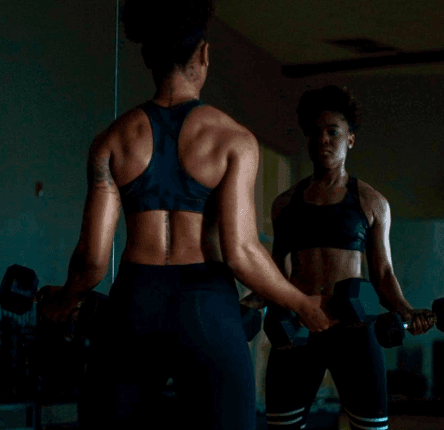 Eine sportlich gekleidete Person hebt Hanteln und betrachtet ihr Spiegelbild in einem Fitnessstudio, wobei ihr muskulöser Rücken und ihre Arme hervorgehoben werden. Die Beleuchtung ist gedämpft und konzentriert sich auf den Körperbau der Person.