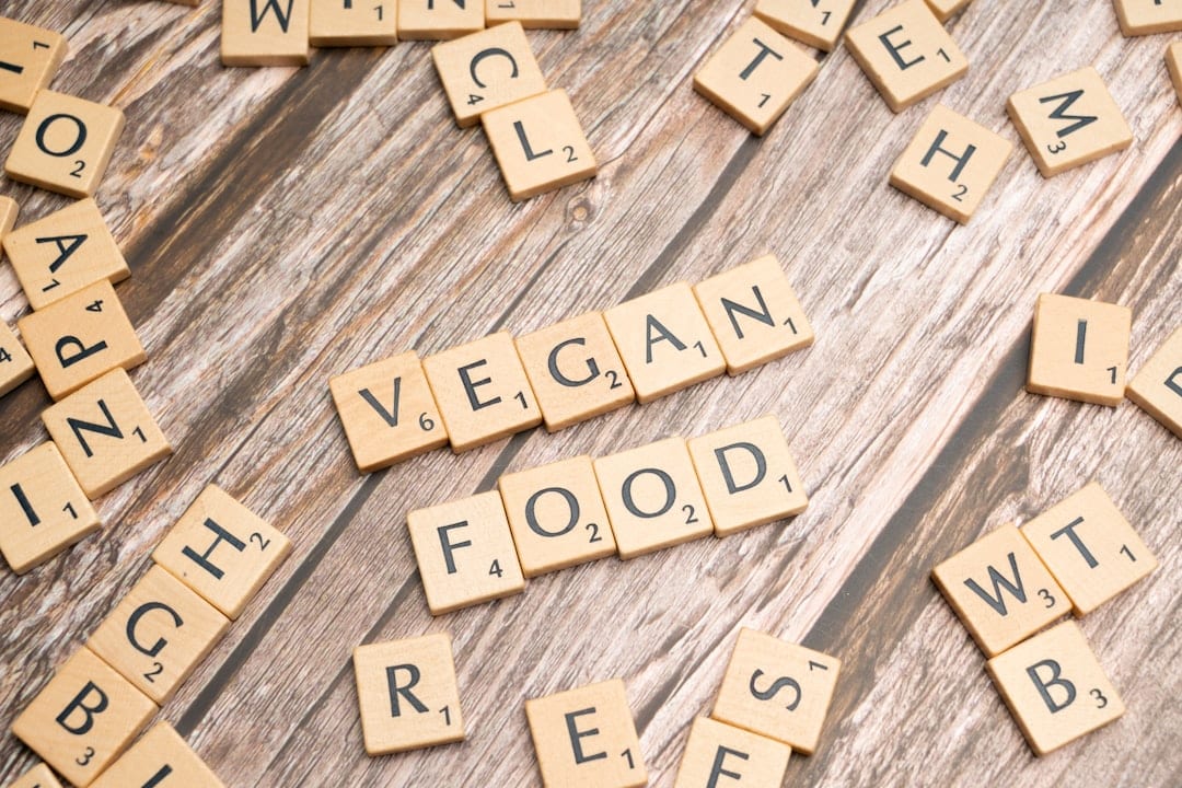 Die Scrabble-Kacheln, die auf einer hölzernen Oberfläche angeordnet sind, ergeben die Buchstaben VEGAN FOOD, die von anderen verstreuten Buchstaben-Kacheln umgeben sind.