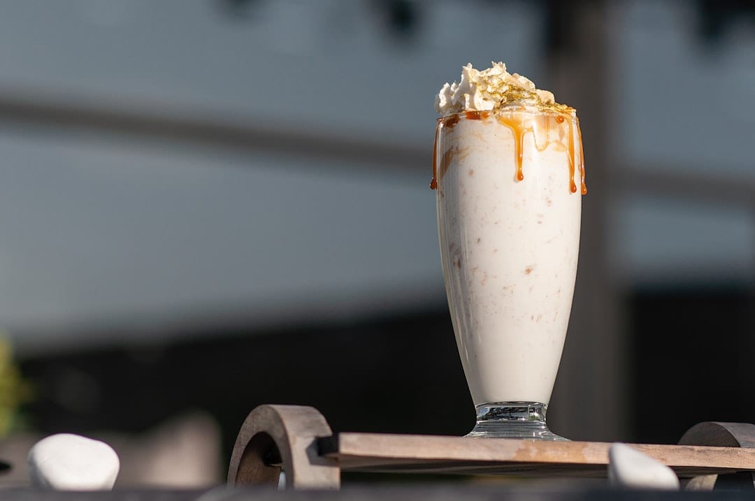 Ein hohes Glas, gefüllt mit einem cremigen Milchshake mit Schlagsahne, Karamellsauce und knusprigen Stückchen, steht auf einer hölzernen Unterlage mit verschwommenem Hintergrund.