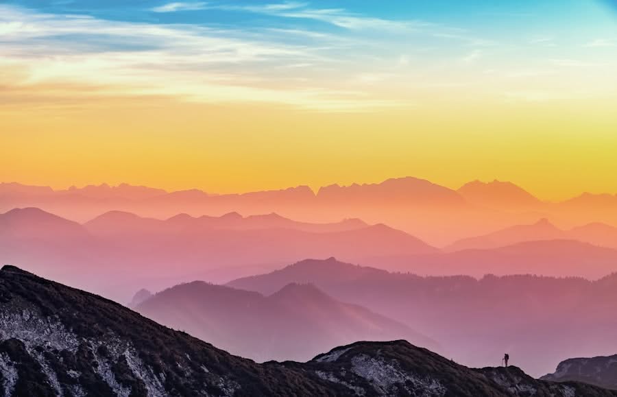 Farbenfroher Sonnenuntergang über einer Bergkette mit orangefarbenen, rosafarbenen und lila Tönen am Himmel. Eine winzige, einsame Figur steht auf einem Hügel im Vordergrund und unterstreicht die Weite der Landschaft.